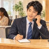 An office employee takes a phone message at a desk while a colleague works nearby