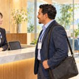 A business visitor checks in at a Japanese office reception desk while a colleague waits to greet him